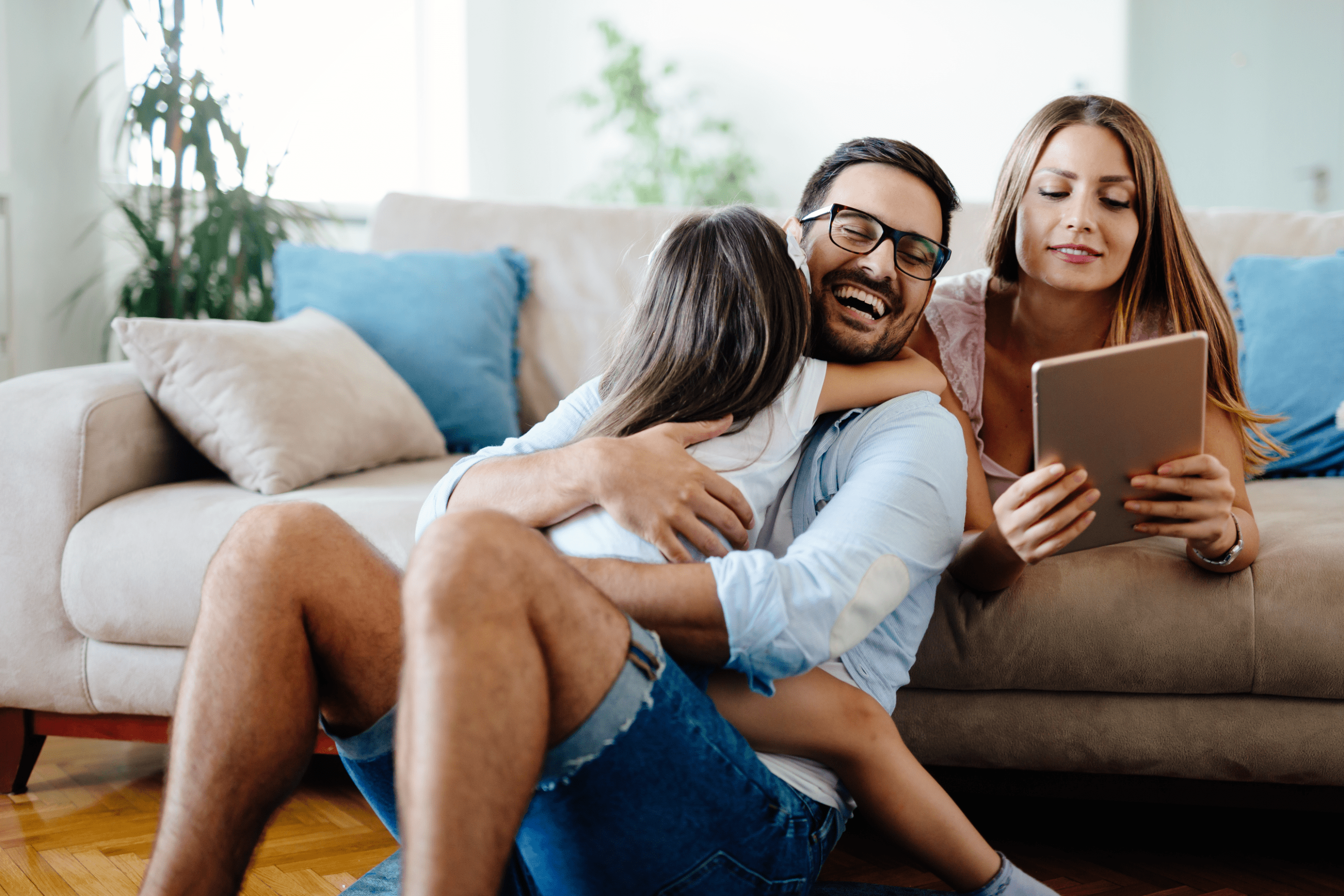 Junge Frau liegt auf einem Sofa und schaut sich etwas auf einem Tablet an. Vor dem Sofa sitzt ein Mann der seine Tochter auf dem Schoß hat. Der Mann lacht. Die Mutter lächelt.